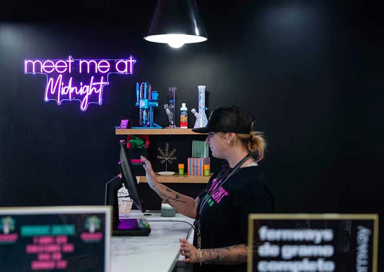 Budtender at Midnight Greens Dispensary assisting a customer under a neon 'meet me at Midnight' sign, reflecting the brand’s modern, women-owned vibe.